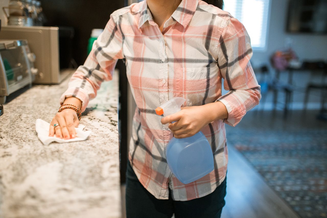 why-choose-us A woman in a plaid shirt wipes down a kitchen counter with a spray bottle and cloth.
